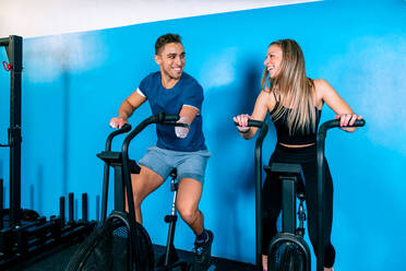 Cheerful young handicapped sportsman and female athlete riding stationary bicycles while looking at each other during functional training in gym - ADSF23086