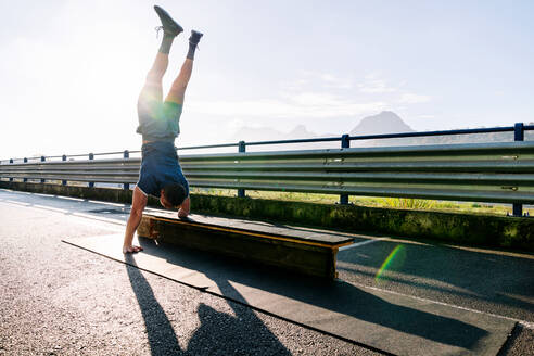 Rückenansicht eines nicht erkennbaren behinderten männlichen Sportlers in Sportkleidung, der auf einer umzäunten Brücke mit Bank im Sonnenlicht steht - ADSF23087