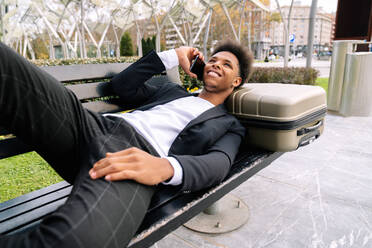 Fröhlicher afroamerikanischer männlicher Reisender, der auf einem Koffer auf einer Bank liegt und mit einem Mobiltelefon spricht - ADSF23089