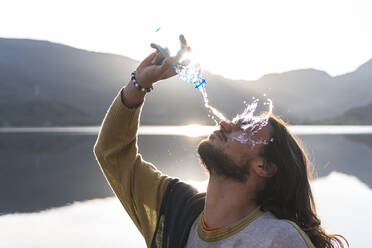 Man pouring water on face with bottle near lake during sunny day - JAQF00504