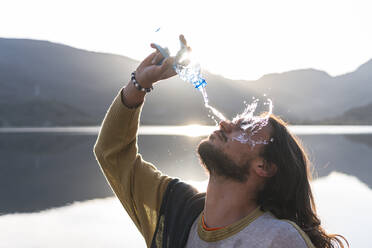 Mann gießt Wasser auf Gesicht mit Flasche in der Nähe von See während sonnigen Tag - JAQF00504