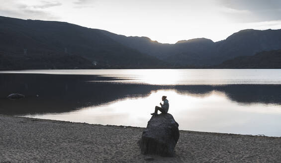 Mann sitzt bei Sonnenuntergang auf einem Felsen am See - JAQF00514