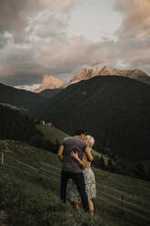 Paar, das sich umarmt, während es im Gras in der Nähe der Dolomitengebirge in Südtirol, Italien, steht - MJRF00397