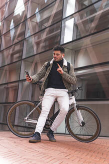 Männlicher Fachmann gestikuliert, während er ein Selfie macht und sich auf ein Fahrrad neben einem Bürogebäude lehnt - JAQF00516