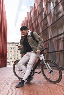 Männlicher Berufstätiger, der sein Smartphone benutzt, während er sich auf dem Gehweg an ein Fahrrad lehnt - JAQF00518