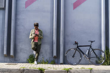 Lächelnder Mann mit Fahrrad, der sein Smartphone benutzt, während er sich auf dem Gehweg an die Wand lehnt - AFVF08615