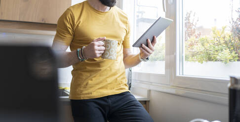Mann, der ein digitales Tablet benutzt, während er eine Tasse zu Hause hält - JCCMF01927