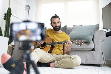 Lächelnder Mann beim Vloggen, während er mit seiner Gitarre zu Hause sitzt - JCCMF01931