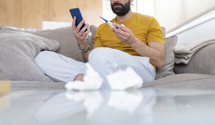 Man holding thermometer and smart phone while sitting on sofa at home - JCCMF01940