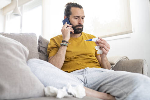 Worried man talking on smart phone while looking at thermometer at home - JCCMF01941