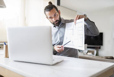 Male design professional showing construction plan on video call over laptop at home - JCCMF01947