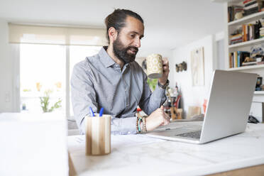 Geschäftsmann mit Tasse sitzt mit Laptop zu Hause - JCCMF01952