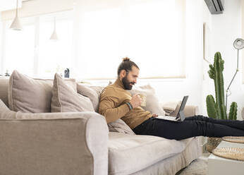 Man with coffee mug using tablet PC while sitting on sofa at home - JCCMF01957