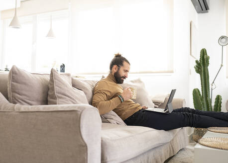 Mann mit Kaffeetasse und Tablet-PC, während er zu Hause auf dem Sofa sitzt - JCCMF01957