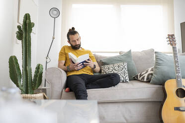 Hübscher Mann liest ein Buch, während er zu Hause auf dem Sofa sitzt - JCCMF01968