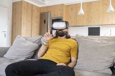 Reifer Mann gestikuliert beim Tragen eines Virtual-Reality-Headsets zu Hause - JCCMF01974