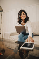 Female freelancer reaching for digital tablet while sitting with laptop at home - DAWF01889