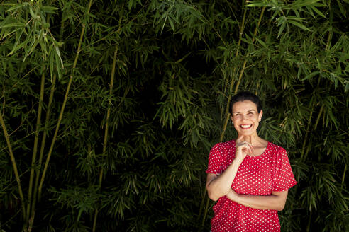 Cheerful woman with hand on chin standing in front of bamboo plants - FLMF00397