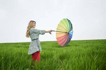 Blond woman playing with multi colored umbrella standing on grass - KIJF03742