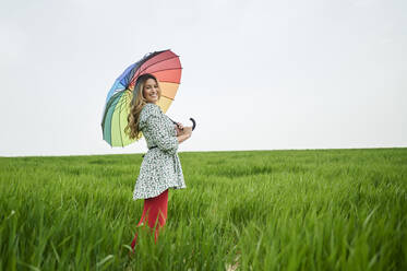 Smiling woman playing with multi colored umbrella standing on grass - KIJF03743