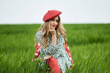 Smiling woman wearing red beret while sitting at meadow - KIJF03745