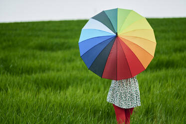 Frau hält bunten Regenschirm, während sie auf einer Wiese steht - KIJF03750