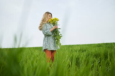 Blonde Frau hält Blumen und steht im Gras - KIJF03767