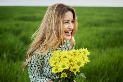 Glückliche Frau schaut weg, während sie gelbe Blumen auf einer Wiese hält - KIJF03768
