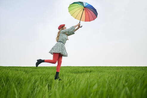 Verspielte Frau mit buntem Regenschirm im Gras auf einer Wiese - KIJF03778