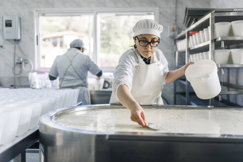 Female chef making cheese with colleague in background at factory - DGOF02098