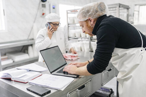 Besitzer benutzt Laptop, während ein Kollege im Hintergrund in der Fabrik arbeitet - DGOF02131