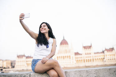 Schöne Frau, die ein Selfie mit ihrem Mobiltelefon macht, während sie auf einer Mauer sitzt, Budapest, Ungarn - ABZF03557