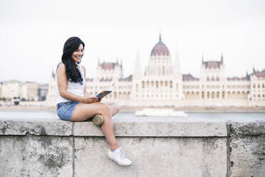 Ein Tourist liest ein Buch, während er auf einer Stützmauer sitzt, mit dem Regierungsgebäude im Hintergrund, Budapest, Ungarn - ABZF03558