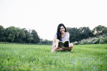 Lächelnde Frau liest ein Buch, während sie im Gras sitzt - ABZF03566