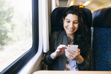 Happy woman using smart phone while sitting in train - ABZF03568