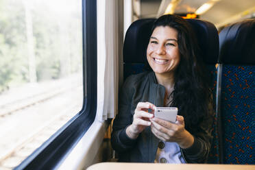 Frau mit Mobiltelefon schaut im Zug sitzend durch das Fenster - ABZF03569