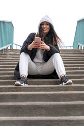 Frau in Kapuzenjacke mit Mobiltelefon auf einer Treppe sitzend - SPCF01337