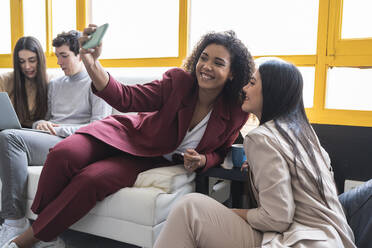Lächelnde Unternehmerinnen machen ein Selfie mit ihrem Smartphone, während ihre Kollegen im Coworking-Büro arbeiten - SNF01228