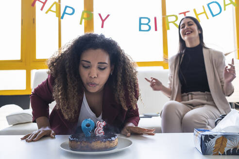 Weibliche Fachkraft bläst Geburtstagskerze auf Kuchen während einer Feier im Coworking-Büro aus - SNF01240