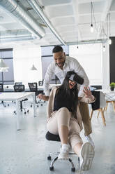 Businessman pushing cheerful female colleague using wearable computer sitting on chair in office - SNF01243
