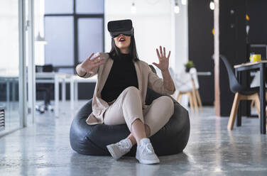 Weibliche Fachkraft gestikuliert, während sie auf einem Sitzsack in einem Coworking-Büro virtuelle Realität genießt - SNF01262