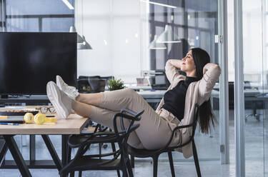 Geschäftsfrau mit Händen hinter dem Kopf entspannt am heißen Schreibtisch im Büro - SNF01267