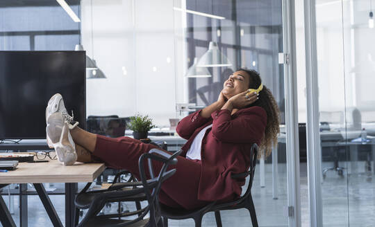 Lächelnde Geschäftsfrau hört Musik über Kopfhörer, während sie mit hochgelegten Füßen auf einem Stuhl im Büro sitzt - SNF01268