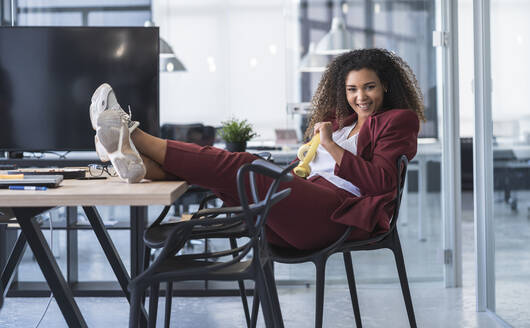 Glückliche weibliche Fachkraft mit hochgelegten Füßen, die sich auf einem Stuhl in einem Coworking-Büro entspannt - SNF01271