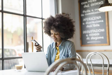 Afro businesswoman with smart phone working on laptop in cafe - EIF00828