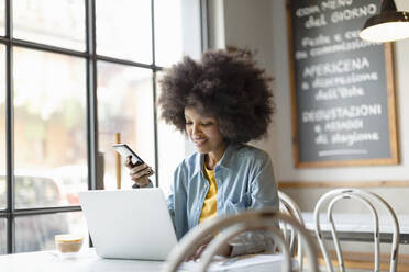 Afro-Geschäftsfrau mit Smartphone arbeitet am Laptop in einem Café - EIF00828