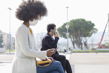 Male and female professionals using smart phones while sitting outdoors during pandemic - EIF00874