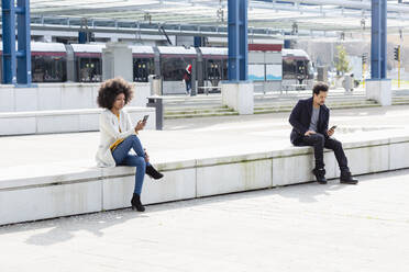 Male and female coworkers using smart phones while following social distancing near railroad station - EIF00876