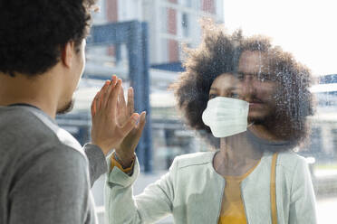 Woman with protective face mask looking at male friend through glass wall - EIF00877