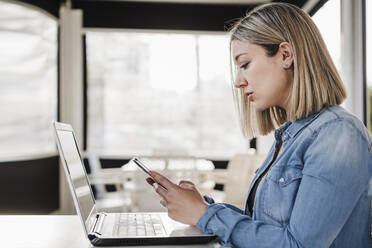 Businesswoman using mobile phone in front of laptop at cafe - EBBF03375